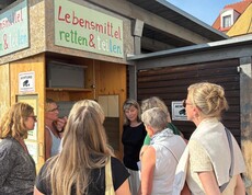 Eine Gruppe Menschen steht vor einem Schrank mit der Aufschrift "Lebensmittel retten und teilen". 