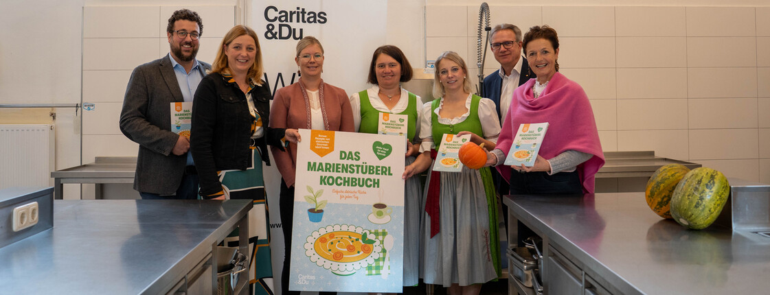 Eine Gruppe von Menschen in der Küche des Caritas Marienstüberls mit einem vergrößerten Schild vom Cover das Marienstüberl Kochbuchs. Von links nach rechts: Marienstüberl-Leiter Adam Lamprecht, Caritasdirektorin Nora Tödtling-Musenbichler, Steirische Seminarbäuerinnen Claudia Kreil, Monika Tatzl und Sandra Hillebrand, SPAR-Geschäftsführer Christoph Holzer und Landwirtschaftskammer-Vizepräsidentin Maria Pein 