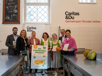 Eine Gruppe von Menschen in der Küche des Caritas Marienstüberls mit einem vergrößerten Schild vom Cover das Marienstüberl Kochbuchs. Von links nach rechts: Marienstüberl-Leiter Adam Lamprecht, Caritasdirektorin Nora Tödtling-Musenbichler, Steirische Seminarbäuerinnen Claudia Kreil, Monika Tatzl und Sandra Hillebrand, SPAR-Geschäftsführer Christoph Holzer und Landwirtschaftskammer-Vizepräsidentin Maria Pein 