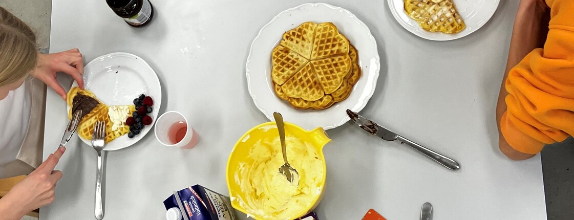 Ein weißer Tisch von oben, gebackene Waffeln und Toppings wie Bananen, Marmelade und Beeren, Teller mit belegten Waffeln und Hände der Mädchen von oben, die hier zusammen sitzen, Waffeln essen und sich unterhalten.