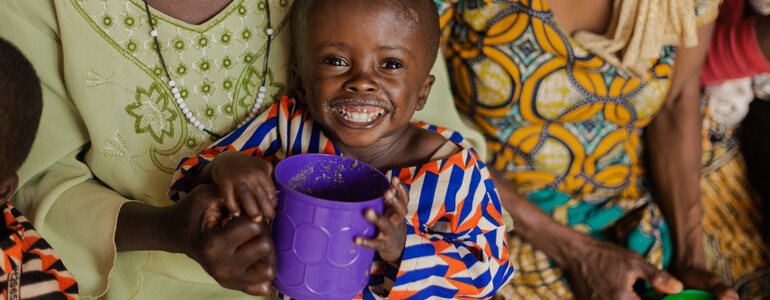 Ein Kleinkind in bunter Kleidung bekommt im Ernährungszentrum in Gitega einen Brei. Es lächelt und schaut zufrieden aus. 