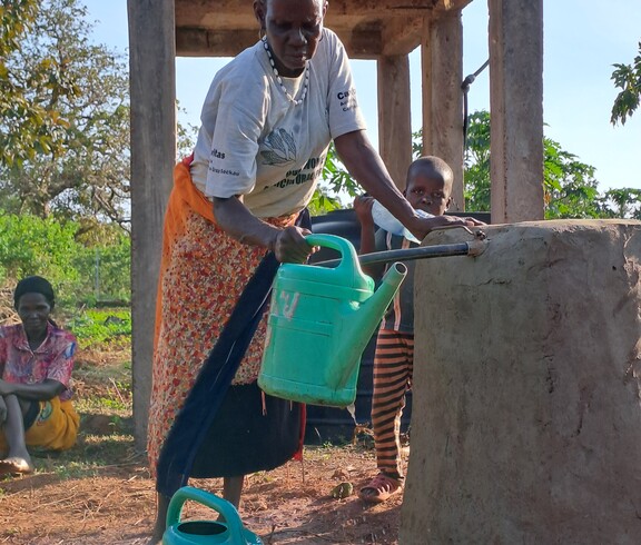 Eine ältere südsudanesische Frau füllt eine Giesskanne an einem Wasserhahn. 