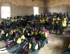 Schulkinder in Schuluniform in einem Lehmzimmer, manche sitzen an Pulten, manche am Boden.