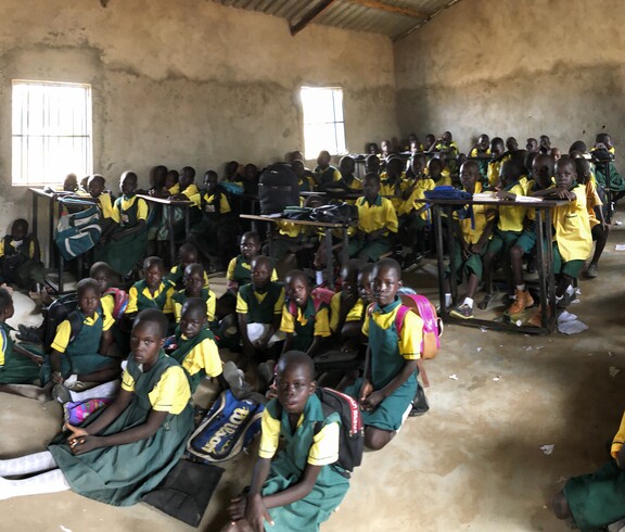 Schulkinder in Schuluniform in einem Lehmzimmer, manche sitzen an Pulten, manche am Boden.