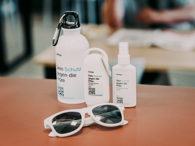 Ein Hitzeschutzpaket, bestehend aus einer Sonnenbrille, einer Trinkflasche mit Karabiner, einer Sonnencreme sowie Insektenschutzmittel auf einem Tisch platziert