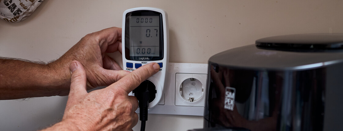 Zwischen Steckdose und Haushaltsgerät ist ein Stromzählgerät angebracht, es wird gerade bedient, ein Knopf wird gedrückt.