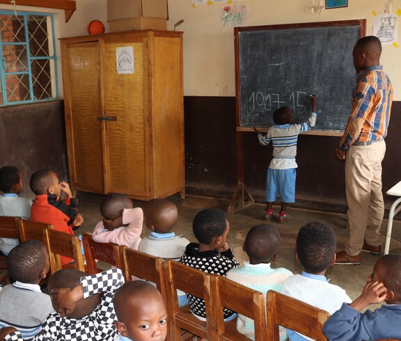 Ein burundisches Kind steht an einer Schultafel und schreibt Zahlen auf. Daneben steht ein Lehrer und man sieht weitere Kinder auf Holzstühlen sitzen. 