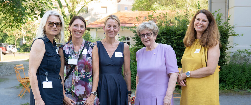 Fünf Frauen mit Namensschildern stehen in einem Garten vor einem Gebäude, eine Frau mit Brille.