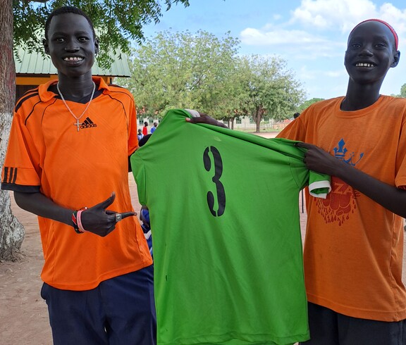 zwei junge Südsudanesen grinsen in die Kamera und halten ein grünes Trikot mit der Nummer 3 zwischen sich. 