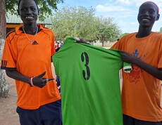 zwei junge Südsudanesen grinsen in die Kamera und halten ein grünes Trikot mit der Nummer 3 zwischen sich. 
