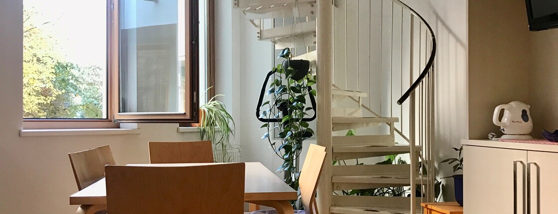 Aufenthaltsraum im Wohnhaus am Volksgarten. Im Raum steht ein Tisch mit Sesseln vor einem geöffneten Fenster mit Blick in den Garten. Im Raum steht auch ein Wandschrank mit cremefarbener Oberfläche. Die Wandfarbe ist weiß, am Fußboden liegt bräunlicher Laminat. Eine Wendeltreppe führt ins darüber liegende Geschoss.  