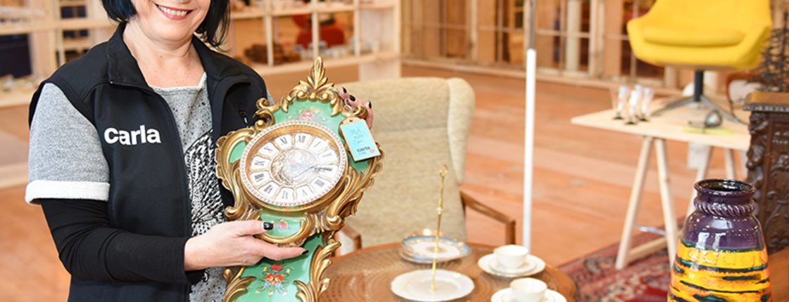 Carla Mitarbeiterin steht vor dekoriertem Tisch mit antiker Uhr im carla Store Bludenz.