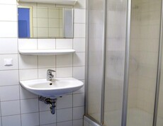 Weißgefliestes Badezimmer mit blauem Boden. Über dem weißen Waschbecken hängt ein Spiegel. Rechts davon befindet sich eine Duschkabine mit Eckeinstieg. Beim Einstieg ist eine Stufe zu überwinden.