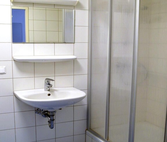 Weißgefliestes Badezimmer mit blauem Boden. Über dem weißen Waschbecken hängt ein Spiegel. Rechts davon befindet sich eine Duschkabine mit Eckeinstieg. Beim Einstieg ist eine Stufe zu überwinden.
