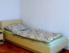 In einer Ecke des Raums steht ein bezogenes Einzelbett. Die Bettwäsche zeigt bordeauxrote Blüten und grüne Ranken. Das Leintuch ist mintgrün. Neben dem Bett steht ein Nachtkästchen mit einer Lade. Die Wandfarbe des Raums ist weiß, der Boden ist Parkett. 