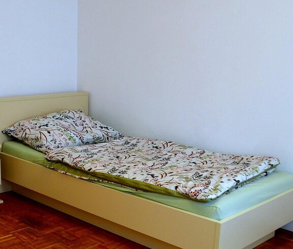 In einer Ecke des Raums steht ein bezogenes Einzelbett. Die Bettwäsche zeigt bordeauxrote Blüten und grüne Ranken. Das Leintuch ist mintgrün. Neben dem Bett steht ein Nachtkästchen mit einer Lade. Die Wandfarbe des Raums ist weiß, der Boden ist Parkett. 