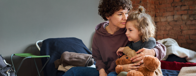 Eine Frau sitzt auf einer Bank, auf ihrem Schoß ein kleines Kind mit Kuscheltier in der Hand.