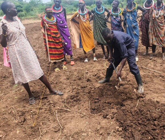 Ein Landwirtschaftlicher Ausbildner zeigt einer Gruppe Frauen, wie sie das Land bearbeiten und ein Beet ausheben. 