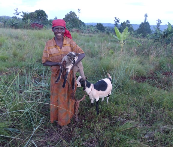 Eine burundische Frau steht lächelnd in einem grünen Feld, eine Ziege an der Leine steht hinter ihr und ein Ziegenjunges trägt sie auf dem Arm. 