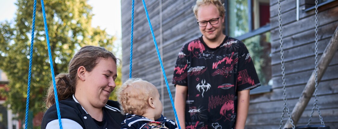 Familienhaus St. Christoph_c_caritas_steiermark  Eine Frau sitzt mit einem Kleinkind auf einer Schaukel, daneben steht ein Mann, der dem Kind zulächelt.