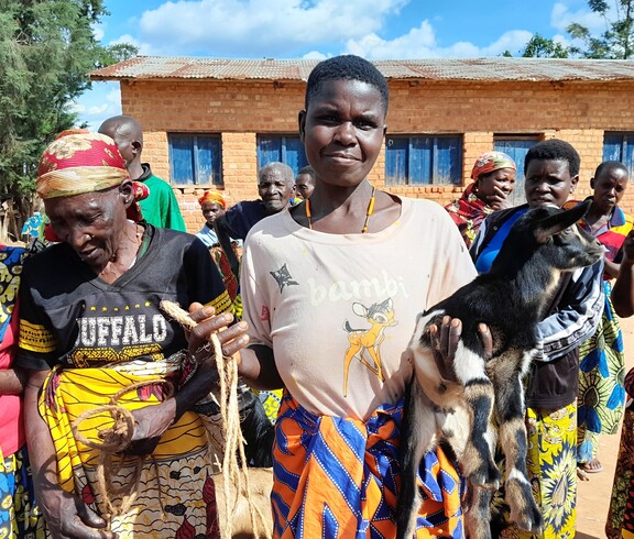 Eine burundische Frau trägt ein Ziegenkitz auf dem Arm und lächelt in die Kamera. Um sie stehen weitere Frauen. 