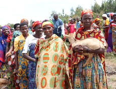 Burundische Frauen stehen aufgereiht in feierlichen Kleidern, die Frau am linken Rand trägt eine Ziege in den Armen. 