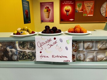 Die "Gesunde Jause"  wurde in der Schule aufbereitet, auf drei Tellern liegt Obst und Gemüse, darunter ein Schild mit gezeichnetem Obst und Gemüse und der Aufschrift "Gesunde Jause, Freie Entnahme"
