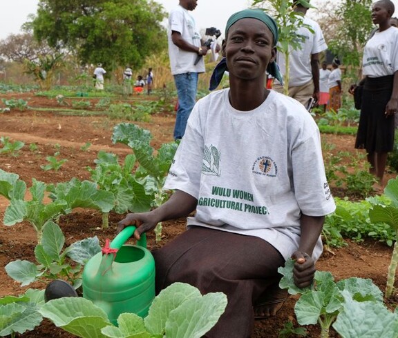 Eine südsudanesische Frau kniet am Boden vor angebautem Gemüse. Eine Giesskanne ist in ihrer Hand.