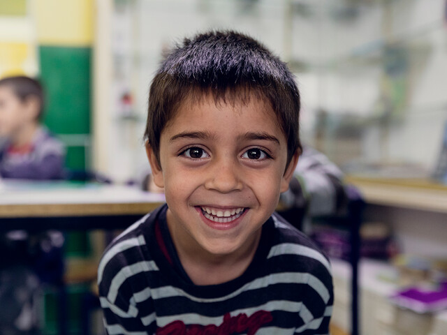 Ein Bub lächelt in die Kamera, im Hintergrund ist verschwommen ein Klassenzimmer zu erkennen.