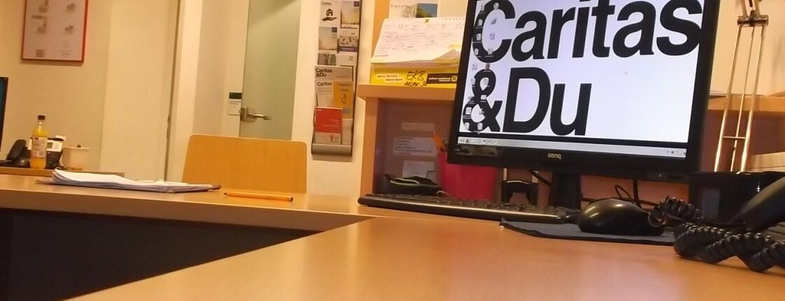 Schreibtisch mit Computer und als Desktop-Hintergrund der Schriftzug "Caritas&Du"