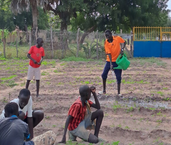 ein paar südsudanesische Burschen sind in einem Garten. Einer gießt, die anderen sitzen am Boden herum. 