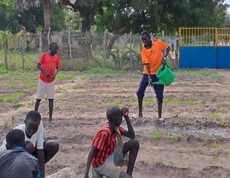ein paar südsudanesische Burschen sind in einem Garten. Einer gießt, die anderen sitzen am Boden herum. 
