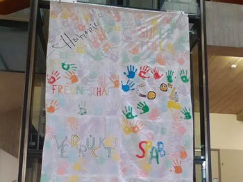 Die Schulflagge ist sehr bunt gestaltet, die Worte Harmonie, Vielfalt, Zusammenhalt, Freundschaft, Verrückt und Spaß sind sichtbar.