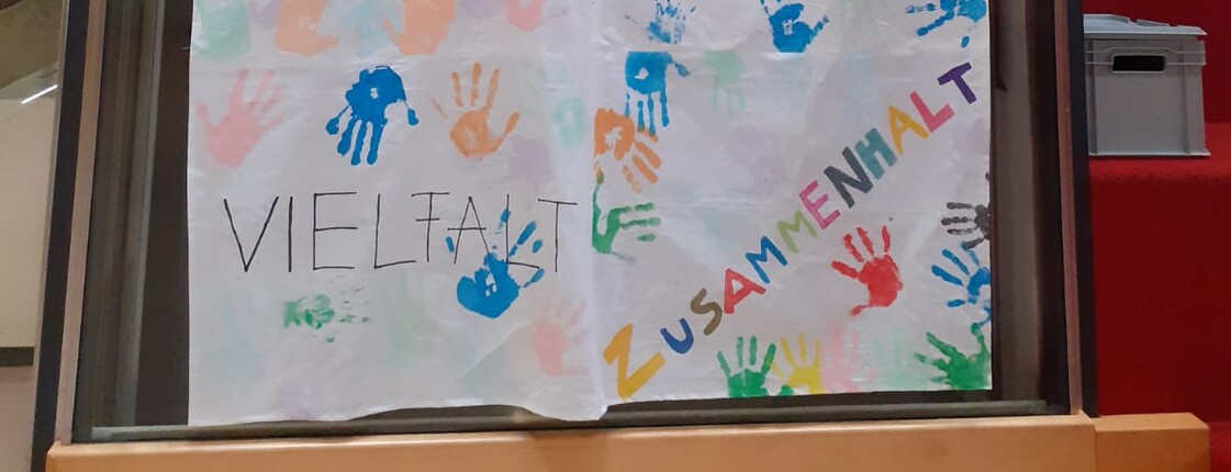 Die Schulflagge ist sehr bunt gestaltet, die Worte Harmonie, Vielfalt, Zusammenhalt, Freundschaft, Verrückt und Spaß sind sichtbar.