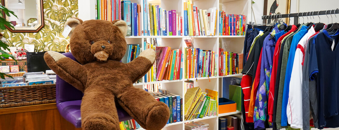 carla Second Hand Shop Ein Bereich des carla Shops im Burgenland mit einem großen braunen Plüschbären, einem Regal voller bunter Bücher und einem Kleiderständer mit Secondhand-Kleidung.