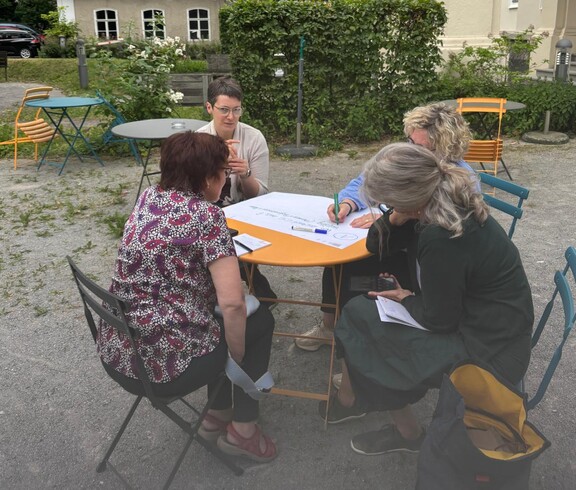 Teilnehmerinnen sitzen um einen Tisch und beschriften ein Plakat. 