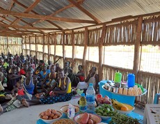 eine Gruppe südsudanesischer Mütter sitzt vor einem blauen Tisch, der mit verschiedenen Zutaten bedeckt ist