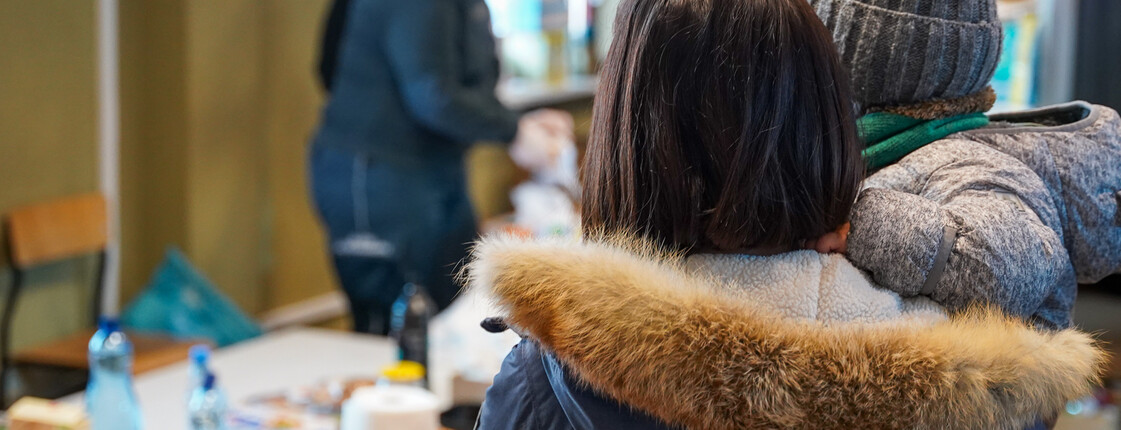 Geflüchtete Mutter mit Kind Eine Frau in Winterkleidung, die ein Kind auf dem Arm hat, steht vor einer Sachspendenausgabe.