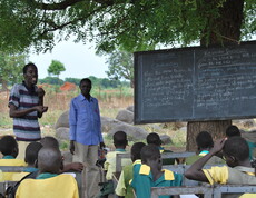 Schüler*innen und Lehrer beim Unterricht unter einem Baum, die Schultafel lehnt an den Stamm des Baumes.