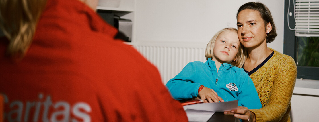 Eine Mutter sitzt mit ihrem Kind auf dem Schoß. Ihnen gegenüber am Tisch sitzt eine Frau mit einer roten Caritas-Jacke, sie ist von hinten zu sehen. 