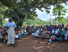 Links steht eine südsudanesische Frau vor einer Gruppe südsudanesischer Frauen, welche in Reihen unter Bäumen sitzen.. 