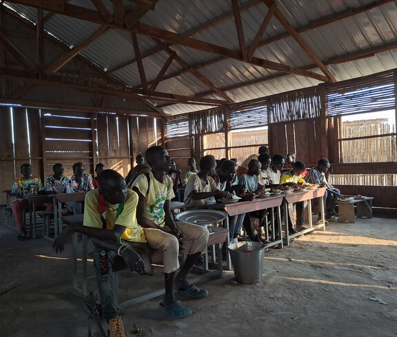 eine Gruppe junger Südsudanesen sitzt in einem düsteren Klassenzimmer und isst. 