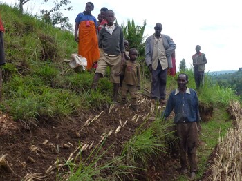 Zwei Männer stehen in einem Gespräch vertieft auf einem begrünten Hang in Burundi; im Hintergrund beobachten weitere Personen die Situation inmitten einer hügeligen, teils bewaldeten Landschaft.