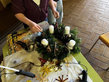 Ein Adventkranz wird auf einem Tisch geschmückt, rundherum liegen Schmuck und eine Heißklebepistole, man sieht die Hände von einer Schülerin, die am Adventkranz arbeitet.