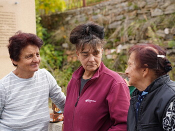 Drei ältere Frauen sind vor einer Steinmauer im Gespräch zu sehen. 