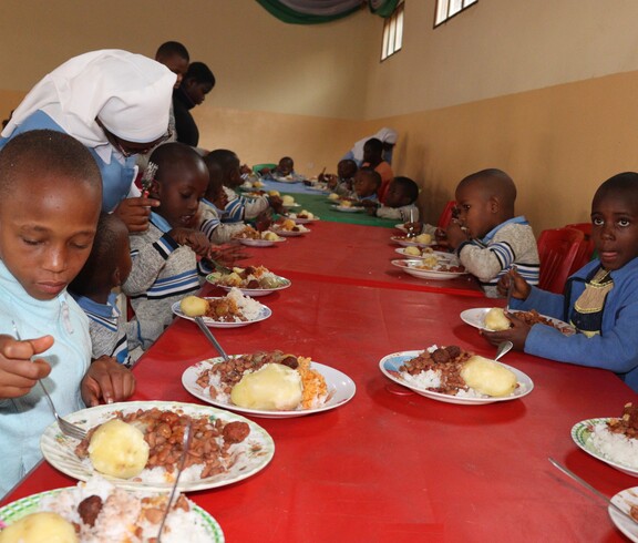 Burundische Kinder sitzen an einem langen Tisch mit rotem Tischtuch und haben einen Teller Essen vor sich. Es gibt Maisbrei und Bohnen. 