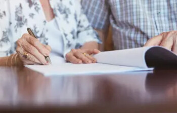 © Canva Ausschnitt zweier älterer Personen, die mit einem geöffneten Dokument auf dem Tisch sitzen.