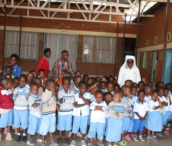 Eine Gruppe burundischer Kindergartenkinder steht mit Blick zur Kamera. Ein paar Ordensschwestern sowie andere Frauen sind im Hintergrund. 