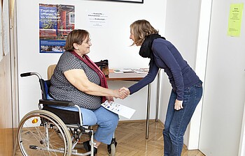 Eine Sozialberaterin begrüßt eine Klientin im Rollstuhl.